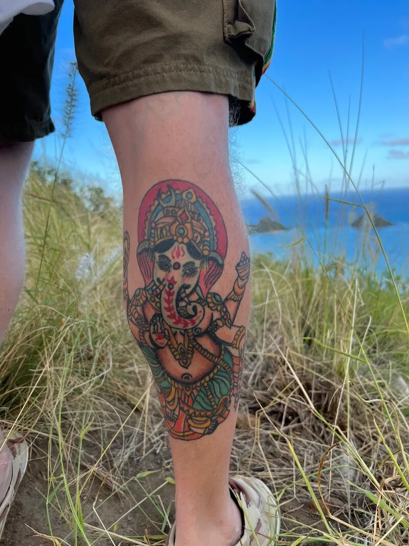 Traditional color tattoo of Lord Ganesha — the Hindu elephant-headed deity — seated in a red halo, on a calf, by Dustin Gormley at Ohana Tattoo, Waikiki, Hawaii.