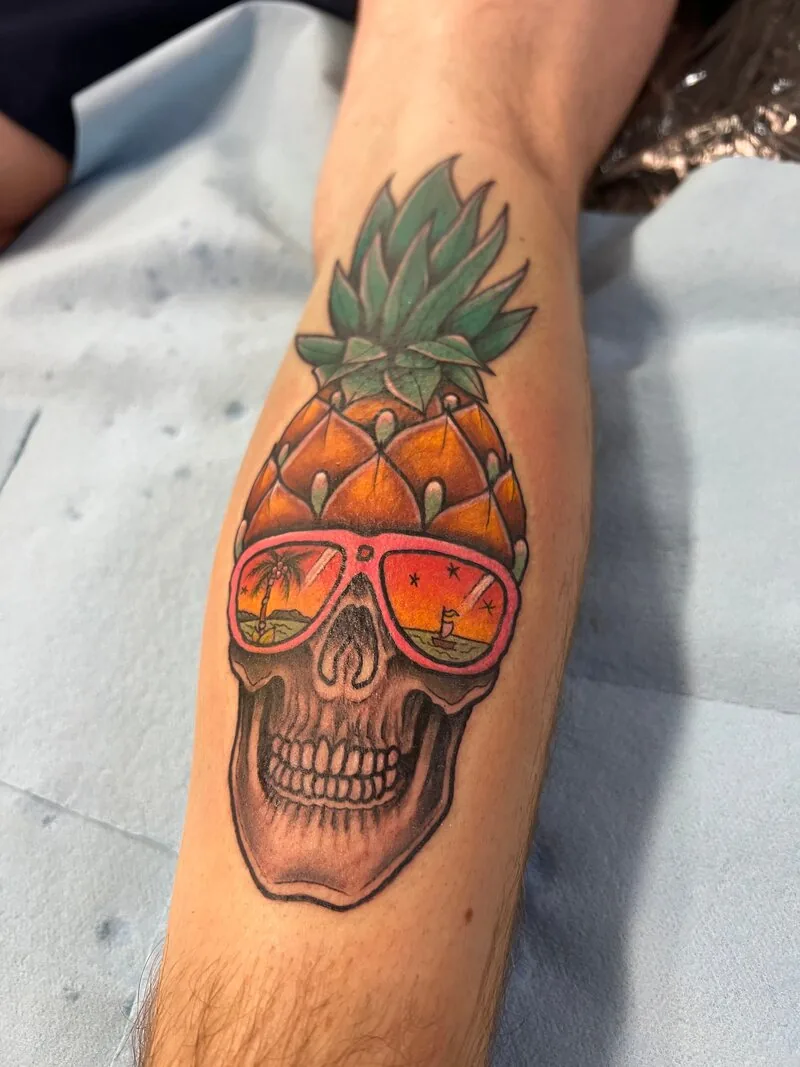 American Traditional tattoo of a pineapple fused with a skull wearing aviator sunglasses, lenses reflecting a Waikiki palm-tree sunset, on a forearm, by Dustin Gormley at Ohana Tattoo, Waikiki, Hawaii.